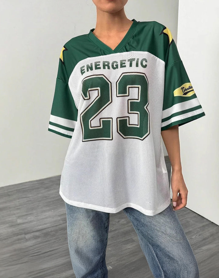 Model wearing a green and white oversized Y2K jersey with 'Energetic 23' print, varsity stripes, and star details. Styled with baggy jeans for a sporty streetwear look.