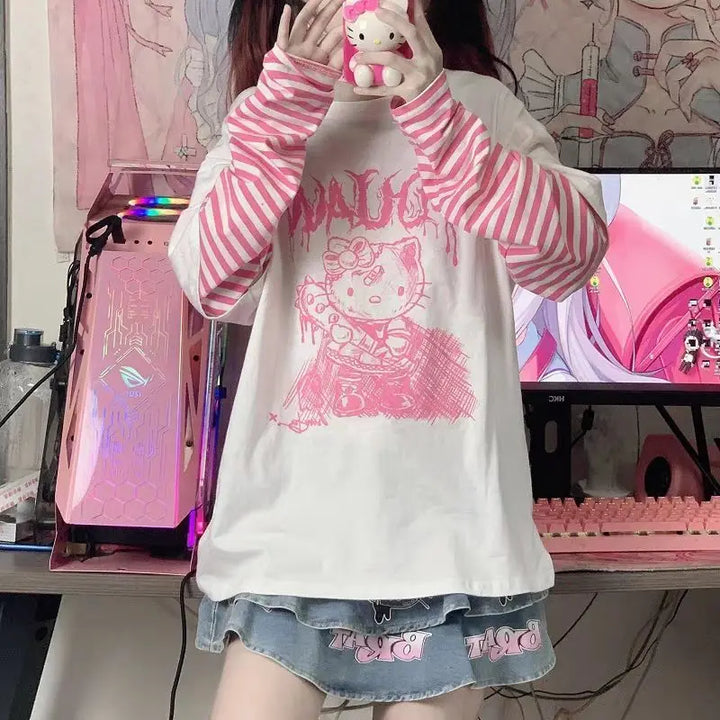 Model wearing a Hello Kitty Harajuku Punk Layered T-Shirt with pink and white striped long sleeves, a denim skirt, and pink leg warmers, posing in a pastel-themed room featuring a pink gaming setup