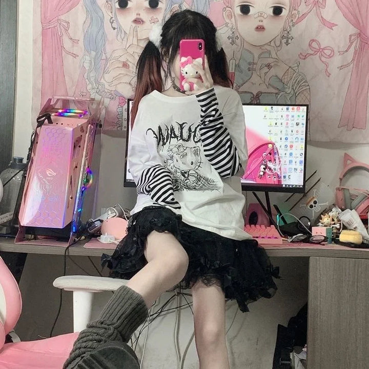 Girl sitting on a desk wearing a Hello Kitty Harajuku Punk Layered T-Shirt with black and white striped long sleeves and a black ruffled skirt, surrounded by kawaii decor and a pastel gaming setup.