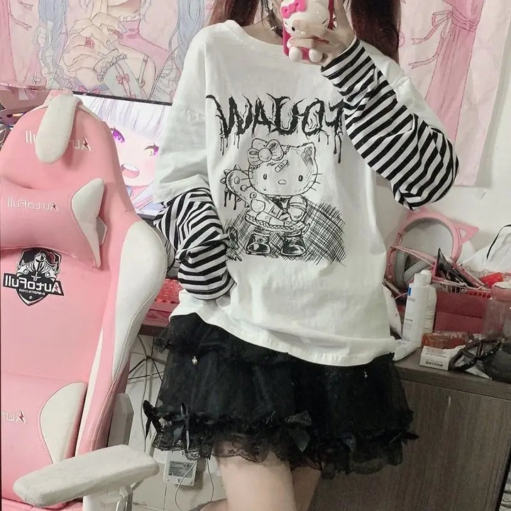 Model dressed in a Hello Kitty Harajuku Punk Layered T-Shirt with black and white striped long sleeves and a black ruffled skirt, standing in front of a pink gaming chair in a kawaii-inspired room.